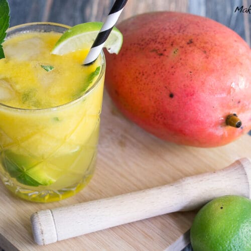 Mojito mit Mango Cocktail 3 Ein Glas Mango-Mojito mit Limette und Minze, daneben eine ganze Mango und Limette auf einem Holzbrett.