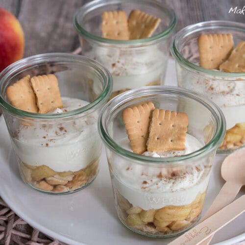 Käsekuchen Dessert mit Apfel 3 Vier Gläser mit geschichtetem Apfel-Käsekuchen-Dessert mit Sahne und Keksen auf einem weißen Teller.