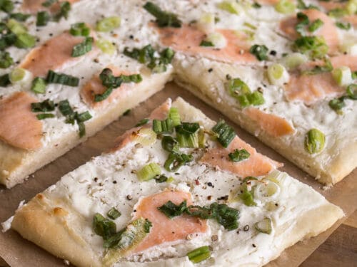 Rechteckiges Flammkuchen, belegt mit Schmand, Räucherlachs, Frühlingszwiebeln und schwarzem Pfeffer auf Backpapier.