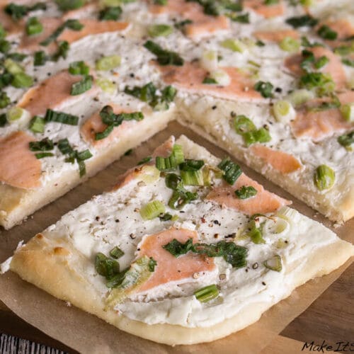 Rechteckiger Flammkuchen mit Räucherlachs mit Schmand und Frühlingszwiebeln, in Quadrate geschnitten auf Backpapier.