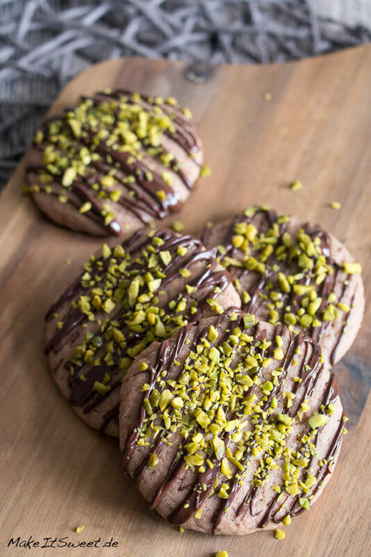Vier Schokoladen-Pistazien-Cookies auf einem Brettchen mit Schokostreifen und gehackten Pistazien verziert