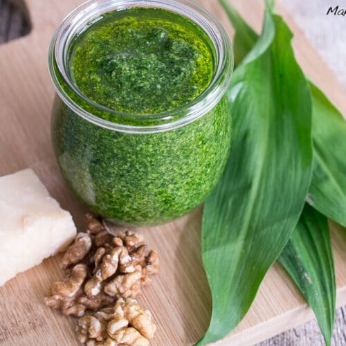 Ein Glas Bärlauchpesto mit Bärlauchblättern, Walnüssen und Parmesan auf einem Holzbrett.