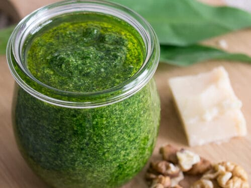 Ein Glas Bärlauchpesto mit Walnüssen, Parmesan und frischen grünen Blättern auf einem Holzbrett.