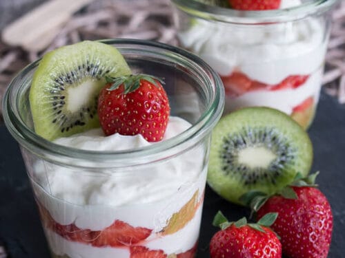 Zwei Gläser Erdbeerdessert mit Kiwi mit geschichtetem Joghurt, Erdbeeren und Kiwischeiben auf einer Schiefertafel.