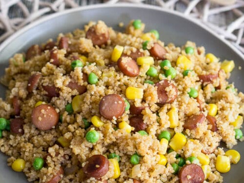 Couscous mit geschnittener Wurst, Mais und Erbsen auf einem grauen Teller aus der Pfanne.
