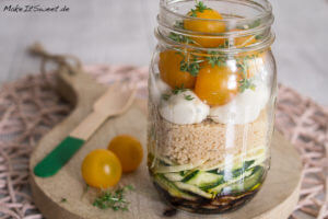Schichtsalat aus gelben Tomaten, Mozzarella, Couscous und Zucchini in einem Einmachglas auf einem Holzbrett.