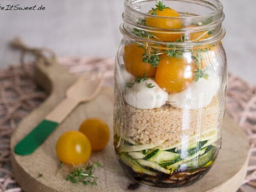 Schichtsalat aus gelben Tomaten, Mozzarella, Couscous und Zucchini in einem Einmachglas auf einem Holzbrett.