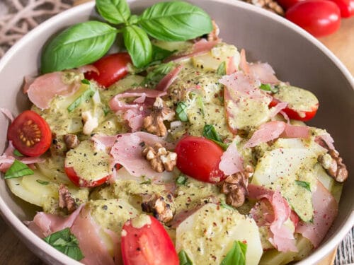 Kartoffelsalat in einer Schüssel mit Tomaten, Prosciutto, Walnüssen, Basilikum und Pesto-Dressing auf einem Holzbrett.