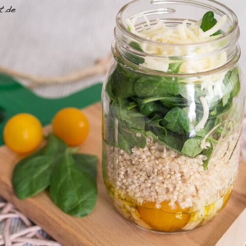 Quinoa Salat mit Tomate und Spinat - Salat im Glas 3 Salat im Glas mit Schichten von gelben Tomaten, Quinoa, Spinat und geriebenem Käse auf einem Holzbrett.