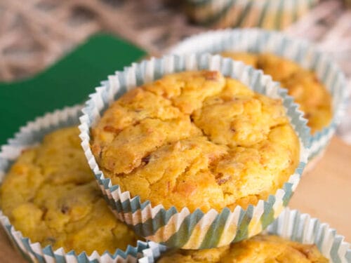 Drei Kürbismuffins mit Käse und Speck auf einem Holzbrett gestapelt, im Hintergrund weitere herzhafte Muffins in gestreiften Beuteln.
