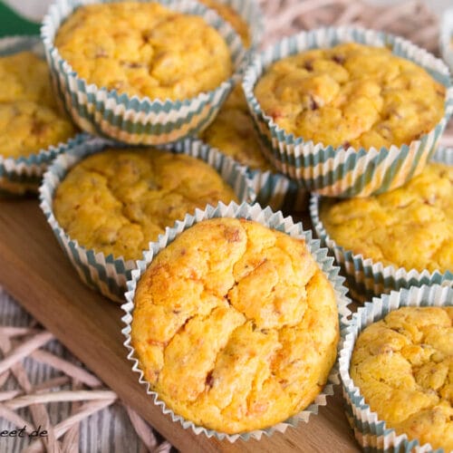 Herzhafte Kürbismuffins mit Käse und Speck 3 Herzhafte Kürbismuffins mit Speck und Käse in gestreiften Förmchen werden auf einem Holzbrett präsentiert.