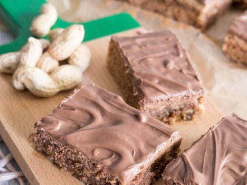 Schokoladenüberzogene Erdnuss-Brownies und geschälte Erdnüsse auf einem Holzbrett.