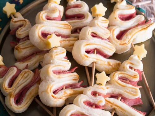 Blätterteig-Tannenbäume mit Schinken am Stiel, garniert mit Käse-Sternen, auf einem Teller serviert.