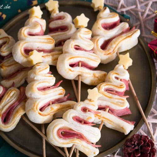 Herzhafte Tannenbäume - Fingerfood zu Weihnachten 4 Blätterteigtannenbäume mit Schinken am Stiel und Käse-Sternen auf einem Teller angerichtet.