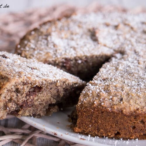 Mehlfreier Nusskuchen mit Puderzucker bestreut, in Scheiben geschnitten auf einem weißen Teller, wobei ein Stück leicht abgetrennt ist.