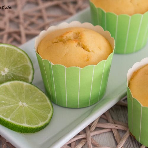 Limetten Muffins mit Joghurt 3 Drei Limetten-Cupcakes in grünen Verpackungen auf einem Teller mit zwei frischen Limettenscheiben.