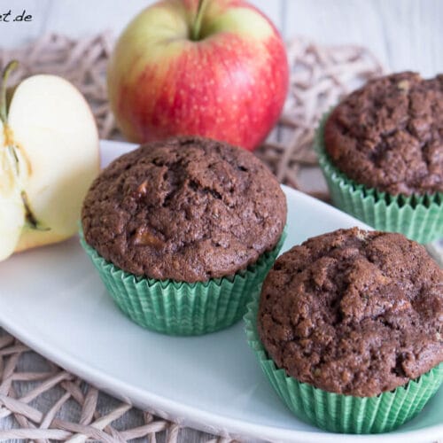 Zucchini Muffins mit Apfel 3 Drei Zucchini-Apfel-Muffins in grüner Verpackung auf einem Teller, dahinter ein ganzer, in Scheiben geschnittener Apfel.