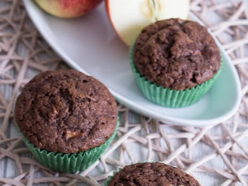 Drei Zucchini-Muffins mit Apfel in grüner Verpackung mit einem ganzen und halbierten Apfel auf einem weißen Teller.