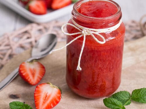 Ein Glas Erdbeermarmelade mit Erdbeeren und Minzblättern auf einem Holzbrett.