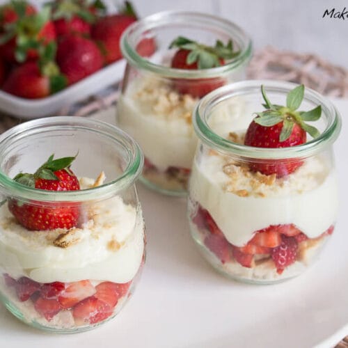 Drei Gläser mit Erdbeeren, Creme und Keksen. Darauf eine Erbessere und zerkleinerte Kekse.