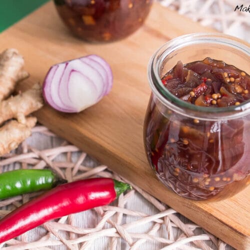 Ein Glas scharfes Zwiebelchutney, daneben Chili, Ingwer und rote Zwiebel, auf einem Schneidebrett aus Holz.