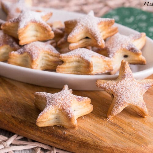 Schnelle gefüllte Weihnachtssterne 3 Mit Puderzucker bestreute sternförmige Kekse, die auf einem weißen Teller und einem Holzbrett angeordnet sind.