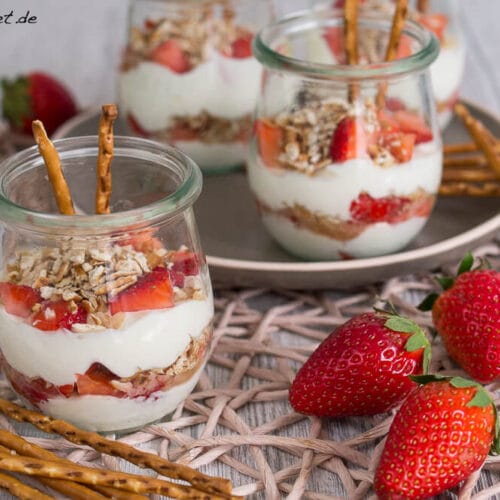 Nachtisch im Glas mit Schichten von Erdbeeren und Laugenstangen, mit zusätzlichen Erdbeeren und Laugenstangen in der Nähe.