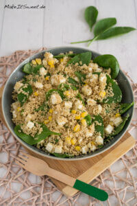 Eine Schüssel Couscous Salat mit Blattspinat, Fetakäsewürfeln und Mais steht auf einem Holzbrett. Im Hintergrund liegen Spinatblätter.