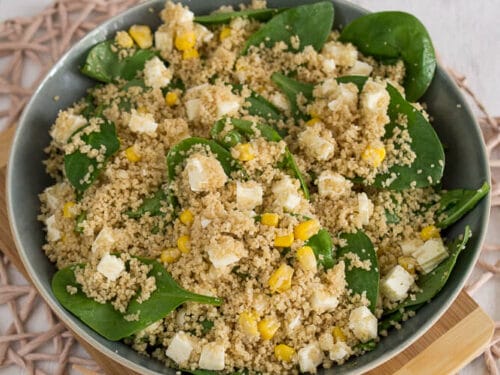 Eine Schüssel Couscous Salat mit Blattspinat, Fetakäsewürfeln und Mais steht auf einem Holzbrett. Im Hintergrund liegen Spinatblätter.