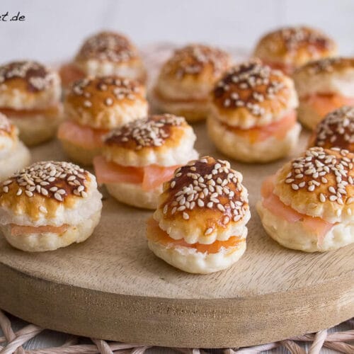Mini-Sandwiches aus Blätterteig, gefüllt mit Räucherlachs und bestreut mit Sesam, angerichtet auf einem Servierbrett aus Holz.
