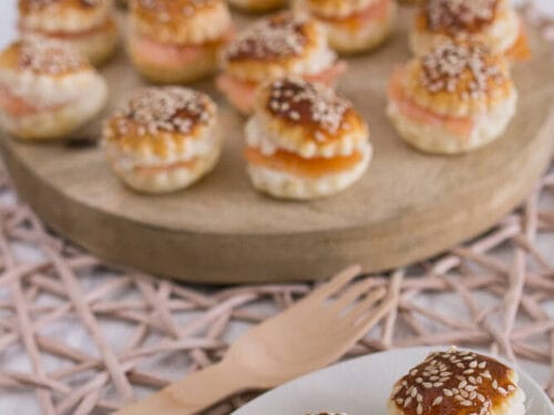 Kleine runde Räucherlachs Burger Blätterteig-Sandwiches mit Sesambelag, gefüllt mit Räucherlachs, sind auf einem Holzbrett im Hintergrund und auf einem kleinen weißen Teller im Vordergrund angeordnet.