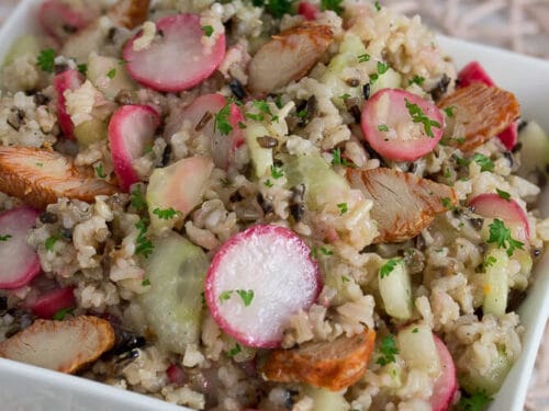 Eine weiße quadratische Schale, gefüllt mit Wildreissalat mit Radieschen, Gurken, Petersilie und gegrillten Hähnchenstücken.