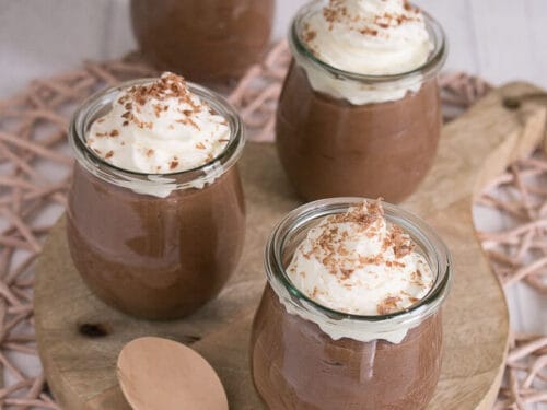 Vier Gläser, gefüllt mit cremiger Nutella Dessert-Schokoladenmousse, gekrönt mit Schlagsahne und Schokoladenraspeln, sind auf einem runden Holzbrett angeordnet. Zwei Holzlöffel liegen vor den Gläsern.