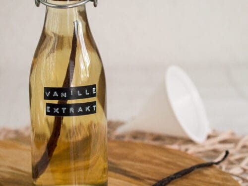 Eine Glasflasche mit der Aufschrift "Vanilleextrakt" und einer Vanilleschote darin steht auf einem Holzbrett. Davor liegt eine aufgeschnittene Vanilleschote, und im Hintergrund ruht ein weißer Trichter.