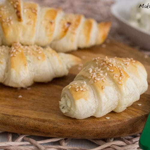 Blätterteig Frischkäse Hörnchen mit Sesam 3 Flockige, goldgelbe Frischkäse-Hörnchen mit Sesam auf einem Holzbrett, serviert mit cremigem Frischkäse.