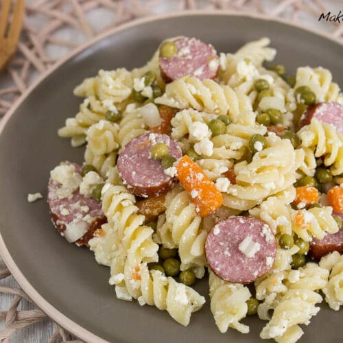 Nudelsalat mit geschnittener Cabanossi, Erbsen, Karotten und zerbröckeltem Feta auf einem grauen Teller.