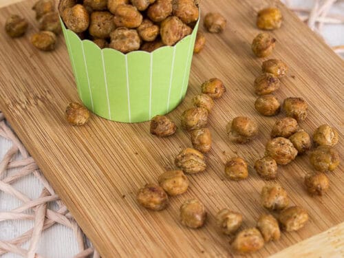 Ein grüner Muffinbecher, gefüllt mit gerösteten Kichererbsen Snack steht auf einem hölzernen Schneidebrett, auf dem weitere Kichererbsen verstreut sind. Das Brett ruht auf einem gewebten Tischset.