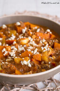 Kürbis-Hackfleisch-Pfanne mit Feta und Kartoffeln 11 Nahaufnahme einer Schüssel mit einer deftigen Kürbis-Hackfleisch-Pfanne mit Kürbisstücken, Kartoffeln, Hackfleisch in einer Tomatensoße und zerbröckeltem Weißkäse. Die Schale steht auf einem gewebten Tischset.