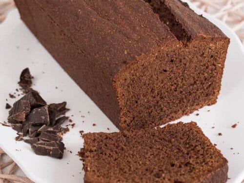 Ein rechteckiger Schokoladenkuchen wird auf einem weißen Teller aufgeschnitten. Es werden zwei Stücke geschnitten, von denen eines flach liegt und das andere aufrecht steht. Mehrere Schokoladenstücke werden neben dem Kuchen platziert.