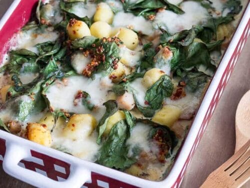 Eine rote Auflaufform mit Gnocchi Auflauf - Gnocchi, geschmolzener Käse, Spinat und körnige Gewürze darauf - steht auf einer Holzplatte neben einem Holzlöffel und einer Gabel.