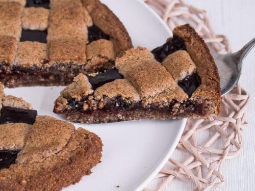 Eine aufgeschnittene Linzer Torte mit Gitterkruste und dunkler Füllung liegt auf einem weißen Teller auf einer gewebten Matte. Eine Scheibe ist leicht angehoben.