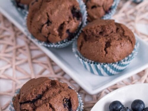 Fünf Schokoladenmuffins mit sichtbaren Blaubeeren liegen auf einem weißen Tellern, ein weiterer Muffin und eine kleine weiße Schale mit frischen Blaubeeren stehen davor auf einem gewebten Tischset.