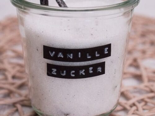 Ein Glas mit der Aufschrift "Vanille Zucker" ist mit selbst gemachtem Vanillezucker gefüllt und enthält eine Vanilleschote. Das Glas ist auf eine gewebte Matte gestellt.