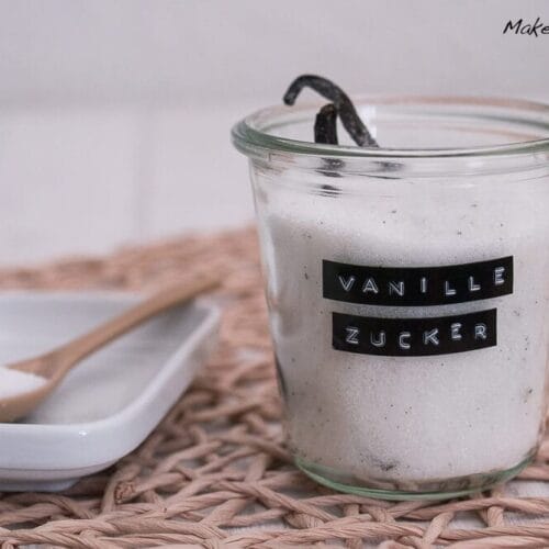Glasgefäß mit hausgemachtem Vanillezucker mit einer Vanilleschote und einem Löffel Zucker, beschriftet mit "Vanille Zucker".