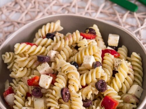 Eine Schüssel griechischer Nudelsalat mit Fetawürfeln, geschnittenen schwarzen Oliven und gewürfelter roter Paprika.