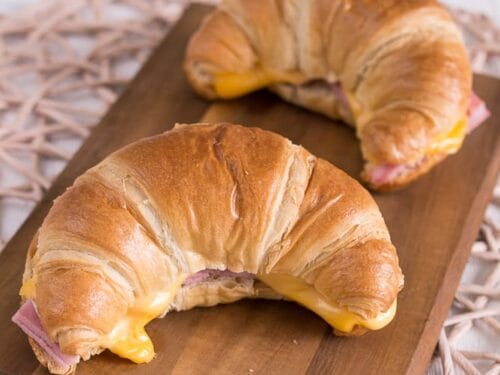 Auf einem hölzernen Servierbrett liegen zwei Croissant mit Schinken und Käse, deren goldene Schichten mit Schinken und geschmolzenem Käse gefüllt sind.