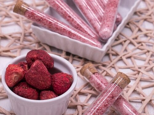 Reagenzgläser mit rosa Erdbeerzucker neben einer Schale mit getrockneten Erdbeeren.