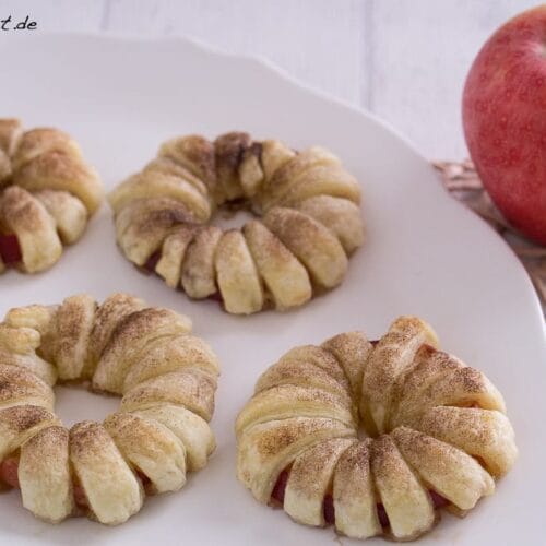Apfel Donuts mit Blätterteig und Zimt 3 Vier Apfel-Donut-Teigringe auf einem weißen Teller neben einem ganzen roten Apfel.
