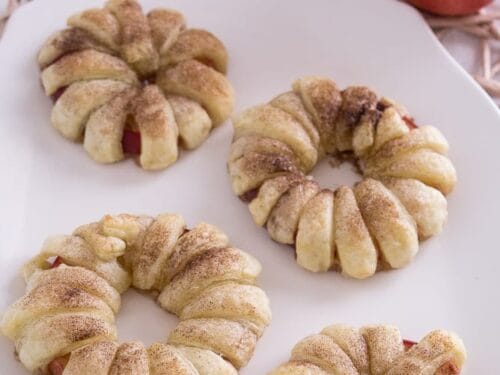 Vier Apfel-Donuts mit Zimtzucker auf einem weißen Teller, daneben ein roter Apfel.