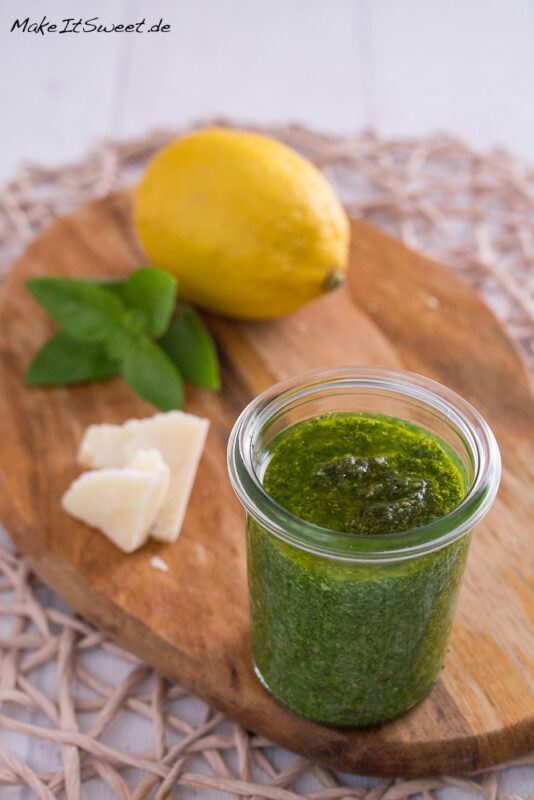 Ein Glas Zitronenpesto mit Basilikum. Die verwendeten Zutaten liegen im Hintergrund